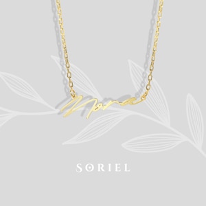 Può includere: Una collana color oro con il nome "Nora" in corsivo. La collana è appesa su uno sfondo grigio chiaro con un sottile motivo a foglie. Il marchio "SORIEL" è visualizzato sotto la collana.