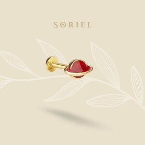 May include: A gold stud earring with a red planet design and a gold band. The earring has a flat back and a post for piercing. The word "SORIEL" is printed in gold letters at the top of the image.