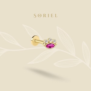 May include: A gold-colored stud earring with a flat back and a decorative front. The front features a marquise-shaped, magenta-colored gemstone, flanked by three smaller, clear gemstones. The word "SORIEL" is printed in gold letters.