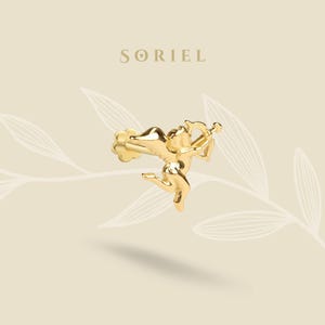 Puede incluir: Un pendiente dorado con forma de Cupido, sosteniendo un arco y una flecha. El pendiente tiene un diseño detallado y está sobre un fondo crema con el nombre de la marca "SORIEL" en la parte superior.