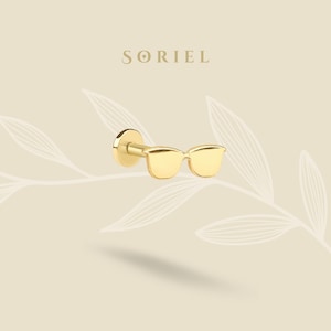 Könnte beinhalten: Ein goldfarbener Ohrstecker in Form einer Sonnenbrille. Der Ohrring hat eine flache Rückseite und einen kleinen Stift. Das Wort "SORIEL" ist in einer Serifenschrift über dem Ohrring aufgedruckt. Der Hintergrund ist hellbeige mit einem Blattmuster.