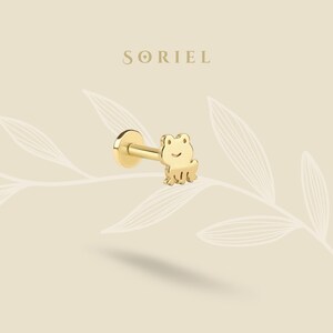 May include: A gold-colored frog-shaped earring with a flat back and a post. The earring has a frog design with a smiling face and small legs. The brand name "SORIEL" is printed in a serif font above the earring.
