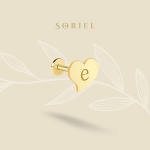 May include: A gold-colored stud earring with a heart-shaped design. The heart features the letter "e" in the center. The earring has a flat back and a post for insertion. The brand name "SORIEL" is displayed above the earring.