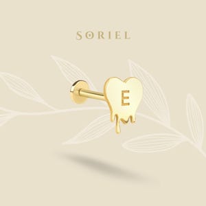 May include: A gold-colored earring with a heart-shaped charm that has a melting design. The letter "E" is in the center of the heart. The earring is on a light beige background with the brand name "SORIEL" at the top.