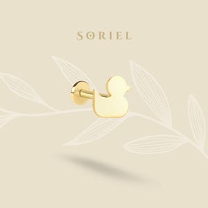 Puede incluir: Un pendiente de botón dorado con forma de pato. El pendiente tiene una parte trasera plana y un poste para la inserción. La palabra "SORIEL" está impresa en una fuente serif en la parte superior de la imagen. El fondo es de color beige claro con un diseño de hojas.