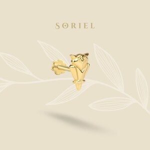 Pode incluir: Um brinco de pressão de coruja dourada. O brinco apresenta um design detalhado de coruja empoleirada em um galho. A palavra "SORIEL" é impressa em letras douradas na parte superior da imagem. O brinco está em um fundo creme com desenhos de folhas.