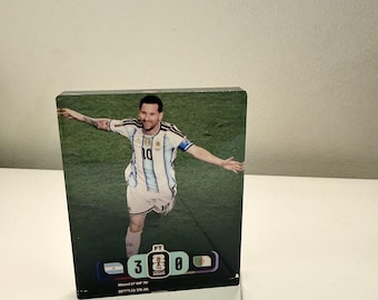 May include: A rectangular acrylic block featuring a printed image of a soccer player in a white and light blue striped jersey with the number 10. Below the player, a scoreboard graphic displays the score 3-0 with the text Messi 17' 60' 70'.
