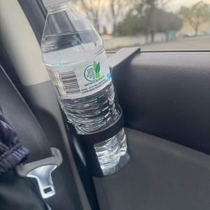 May include: A black plastic car cup holder with a clear plastic water bottle inside. The bottle has a green label with the text "Sea of Bottle" and "Droppy Bottles and Caps".