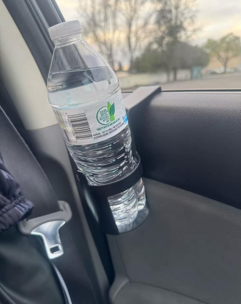 May include: A black plastic car cup holder with a clear plastic water bottle inside. The bottle has a green label with the text "Sea of Bottle" and "Droppy Bottles and Caps".
