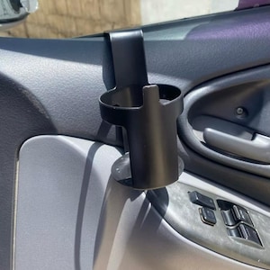 Peut inclure: Porte-gobelet de voiture noir monté à l'intérieur d'une portière de voiture. Le porte-gobelet est en métal et a une finition noire.