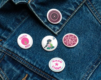 Könnte beinhalten: Eine Sammlung von fünf runden Buttons mit verschiedenen Designs. Ein Button zeigt ein rosa Mandala, ein anderer ein Mädchen mit einem Buch, und ein dritter Button sagt "Kosmic Girl" mit einer Gitarre. Die anderen beiden Buttons haben einen Stern und die Worte "Moon Light".