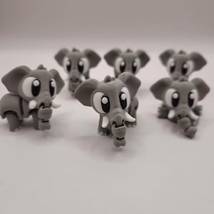 Op de afbeelding: Een verzameling van zes kleine, grijze, 3D-geprinte olifantfiguurtjes. Elke olifant heeft grote, wijd uit elkaar staande zwart-witte ogen, opvallende oren en kleine witte slagtanden. De gelaagde textuur van het printproces is zichtbaar.