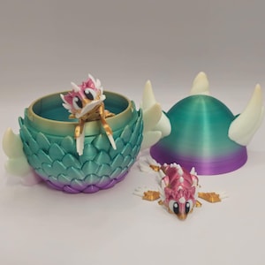 Puede incluir: Un conjunto temático de dragones impreso en 3D. El conjunto incluye un recipiente verde azulado, morado y dorado con un dragón, una figura de dragón y un casco verde azulado y morado con cuernos blancos. Las figuras de dragón tienen detalles rosas y dorados.