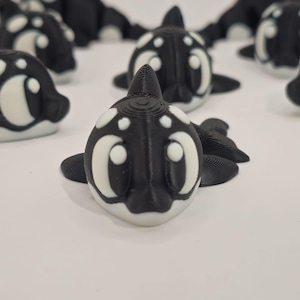 May include: A collection of black and white orca figurines. Each figurine has a detailed design, with a black body and white accents around the eyes and on the body. The figurines are arranged on a white surface.