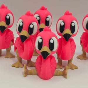 Peut inclure: Un groupe de cinq petites figurines de flamants roses imprimées en 3D. Chaque figurine présente de grands yeux blancs avec des pupilles noires, un bec beige à pointe noire et des pattes beiges. Le flamant central est assis.