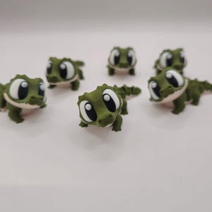 Op de afbeelding: Een verzameling van zes kleine olijfgroene alligatorfiguurtjes. Elk figuurtje heeft een getextureerd, geschubd lichaam, een lichtgekleurde onderbuik en extra grote, cartoonachtige witte ogen met grote zwarte pupillen, verspreid opgesteld.