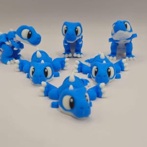 Op de afbeelding: Een verzameling blauwe en witte 3D-geprinte drakenfiguren. De draken hebben grote ogen, witte klauwen en kleine hoorns. Sommige staan, andere liggen, wat een speelse en grillige weergave creëert.