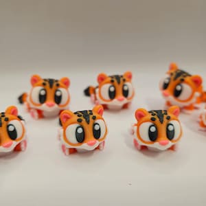 Può includere: Una collezione di piccole statuette di tigre arancioni e bianche con strisce nere e grandi occhi da cartone animato. Le statuette hanno i piedi rosa e sono disposte su una superficie bianca.
