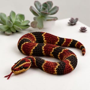 Puede incluir: Un juguete de serpiente tejido a ganchillo con un patrón repetitivo de bandas de color rojo oscuro, negro y amarillo. La serpiente tiene una cabeza redondeada con pequeños ojos negros y una lengua roja fina y bífida que sale de su boca.