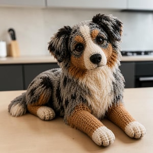 May include: A crocheted stuffed animal in the likeness of an Australian Shepherd dog. The plush features a multi-coloured coat of grey, black, white, and tan yarn, with dark brown eyes, a black nose, and a textured, shaggy appearance throughout.