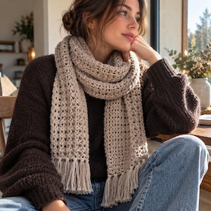 Puede incluir: Una bufanda larga de ganchillo en color beige con un patrón de punto calado y terminada con borlas de flecos rectos en ambos extremos. La bufanda está envuelta alrededor del cuello, mostrando la textura del hilo y su tono neutro.