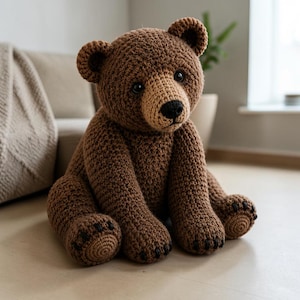 May include: A brown crocheted teddy bear with a lighter tan muzzle, dark round eyes, and a black stitched nose. The bear has textured yarn construction, rounded ears, and dark stitched details on the paws, sitting in an upright position.