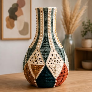 May include: A decorative vase covered in a multi-colored crochet sleeve featuring geometric triangular patterns in shades of dark green, cream, mustard yellow, and terracotta. The textured knit design includes raised bobble stitches.