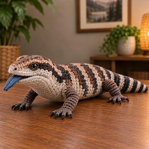 Puede incluir: Un juguete de peluche de lagarto tejido a ganchillo con un cuerpo texturizado de rayas alternas en marrón, beige y crema. El lagarto tiene una lengua azul prominente que sobresale de la boca, ojos marrón oscuro y cuatro patas con garras negras.