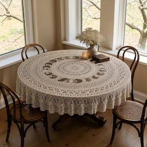 May include: A round, beige crochet tablecloth featuring a central mandala pattern surrounded by a lunar phase motif. The intricate lace fabric drapes over a circular table, which is accompanied by four dark wood chairs with woven seats.