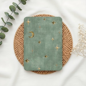 May include: A sage green rectangular card featuring a celestial design. Gold stars and a crescent moon are scattered across the card's surface. The card is placed on a woven, circular placemat, with eucalyptus and white flowers in the background.