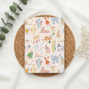 May include: A cream-colored notebook featuring a whimsical pattern of illustrated cats in various poses and outfits. The cats are adorned with floral patterns, hats, and accessories. The notebook is placed on a round, woven wicker placemat.