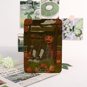 May include: A rectangular card featuring a fall-themed illustration. A figure with a pumpkin head sits in a shopping trolley in front of a sign that reads "Pumpkin Patch". Pumpkins are scattered around the trolley. Other cards and decorative elements are in the background.