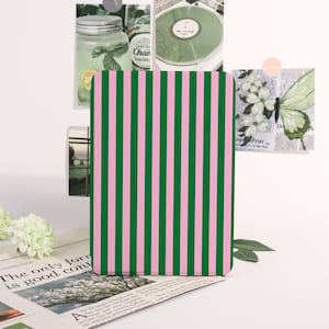 May include: A notebook featuring a pink and green striped design. The notebook is positioned on a newspaper, with images of a jar, a record, and a butterfly in the background.
