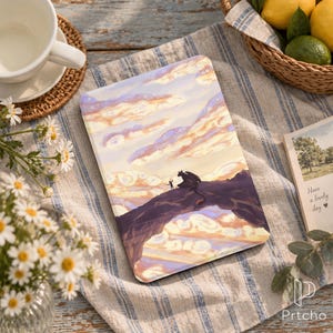 May include: A rectangular e-reader case featuring a printed illustration of a person standing on a dark rock formation with a dragon against a pastel sunset sky with swirling clouds. The case is displayed on a blue and white striped fabric surface.