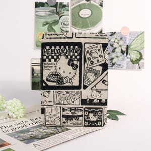 May include: A Hello Kitty themed notebook with a black and white comic strip design. The cover features the text "HELLO KITTY" and illustrations of the character. The notebook is displayed with other decorative items.