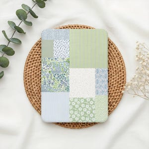 May include: A rectangular notebook with a patchwork design featuring various patterns in shades of green, blue, and white. The notebook is placed on a round woven placemat, with eucalyptus and white flowers in the background.