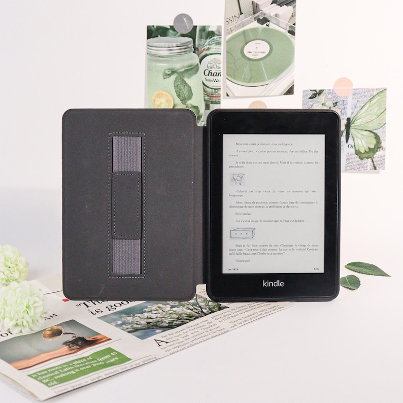 May include: A black Kindle e-reader with a dark gray cover, open to display text. The cover has a gray elastic hand strap. The Kindle is on a white surface with a newspaper and decorative items in the background.