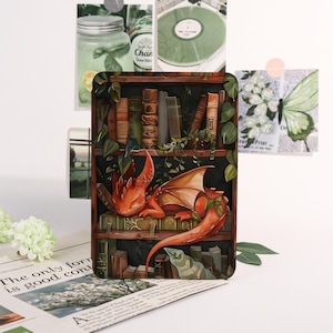 May include: A rectangular decorative item with an illustration of a red dragon on a bookshelf. The dragon has orange wings and is surrounded by green vines. The bookshelf is filled with books. Other decorative elements are in the background.