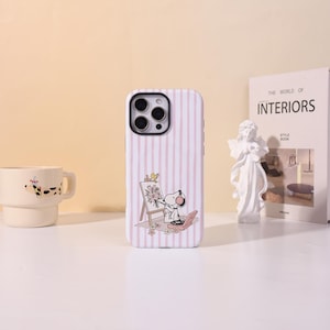 May include: A light pink and white striped phone case featuring a graphic of Snoopy wearing headphones while painting a bouquet of flowers on an easel, with a small bird perched on top and a record player placed on the ground near the easel.
