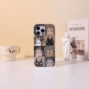 May include: A phone case featuring a collage of cat faces in various styles and colors. The case has a rectangular shape with rounded corners and a camera cutout. The cat illustrations include black, white, and tan cats with different expressions and patterns.