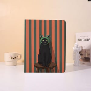 May include: A black cat wearing a green frog hat sits on a small wooden stool in front of a striped orange and green background. The cat is the focal point, with a whimsical and playful design. A white mug with a dog design is on the left.