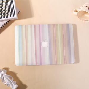 Op de afbeelding: Een rechthoekige harde laptophoes met een verticaal streeppatroon in zachte pasteltinten van roze, blauw, groen en lavendel. De hoes heeft een centrale uitsparing die het zilveren Apple-logo onthult en een gladde, semi-transparante afwerking.
