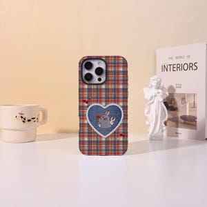 May include: A rectangular smartphone case featuring a multi-colored plaid pattern in shades of red, blue, yellow, and white. A central denim-textured heart patch displays a cartoon rabbit holding a red flower, accented by small cherry illustrations.