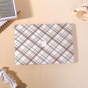May include: A laptop with a white and gray plaid pattern. The Apple logo is visible in the center. The laptop is on a light beige surface, with a stack of books and a mug in the background.