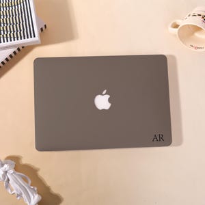 Op de afbeelding: Een gesloten grijze laptop met het Apple-logo in het midden en de initialen "AR" rechtsonder. Een witte mok met een bloemenpatroon en een stapel boeken zijn ook zichtbaar.
