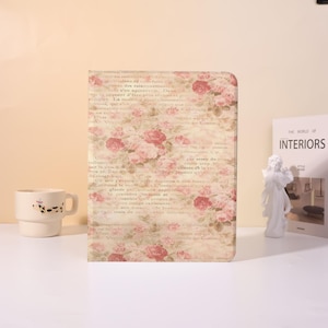 May include: A notebook with a floral design, showcasing a vintage style. The cover displays a faded backdrop with pink roses and script-like text. A white mug and a book titled "The World of Interiors" are also present.