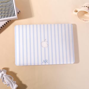 May include: A light blue and white striped laptop case with a cut-out apple logo. The initials "AR" are printed in navy blue at the bottom. A white coffee mug with a floral design and a stack of books are in the background.