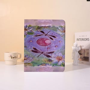 May include: A journal with a watercolor painting of dragonflies over a pond with lily pads. The cover art features a purple and pink color palette with a surrealist style. A white mug and a book are in the background.