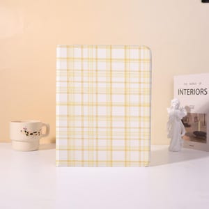 May include: A white and yellow plaid pattern notebook stands upright on a white surface. A ceramic mug with a black and white pattern and a decorative statue are to the left and right of the notebook. A book titled "INTERIORS" is also visible.