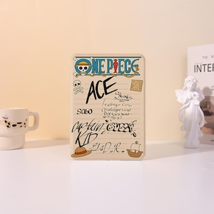 May include: A rectangular beige tablet case featuring the One Piece logo at the top, followed by various handwritten-style names and phrases including Ace, Sabo, Captain Kid, and Buggy, along with small illustrations of a straw hat and a ship.
